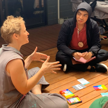 Group of four people sitting on a wooden deck engaged in discussion, with colourful cards and papers spread on the floor.